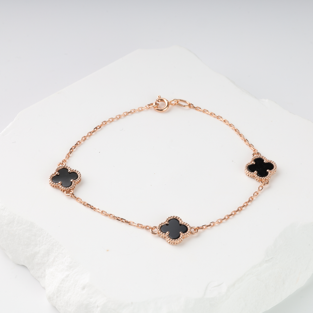 Rose gold chain bracelet with three black enamel four-leaf clover charms displayed on a white stone surface.