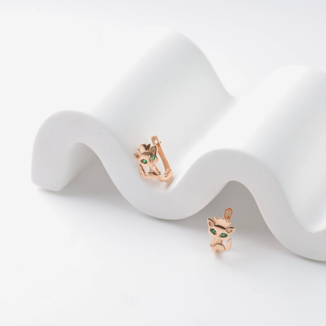 Rose gold cat earrings with green gemstones displayed on a white wavy stand.