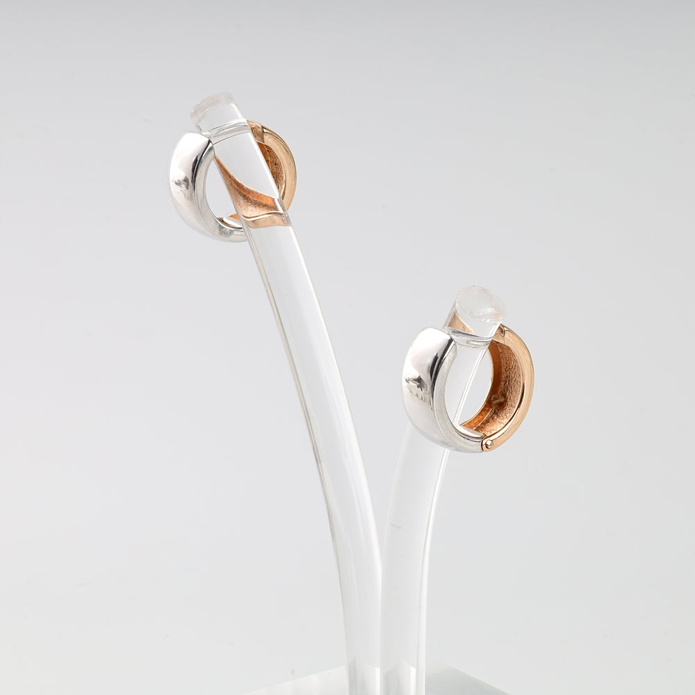 Pair of silver and rose gold huggie hoop earrings displayed on a clear acrylic stand.