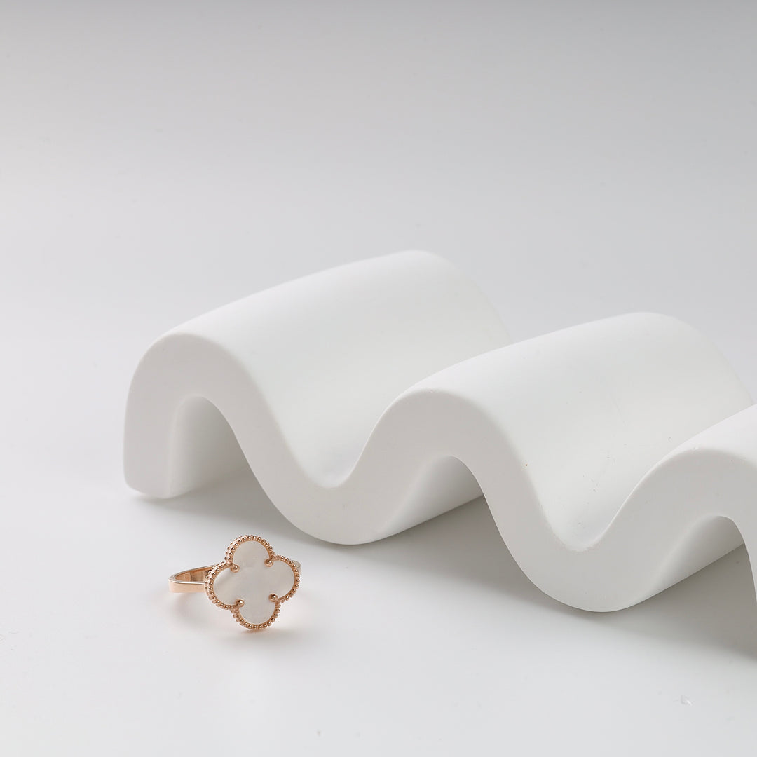 Gold ring with a white clover-shaped stone on a curved white display, minimalist jewelry photo.