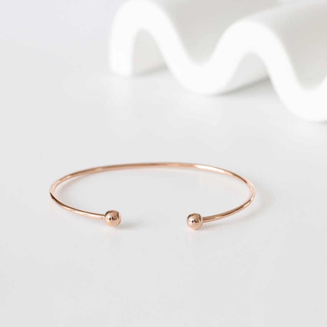 Rose gold open cuff bracelet with round ball ends sits on a white surface, minimalist jewelry bangle.