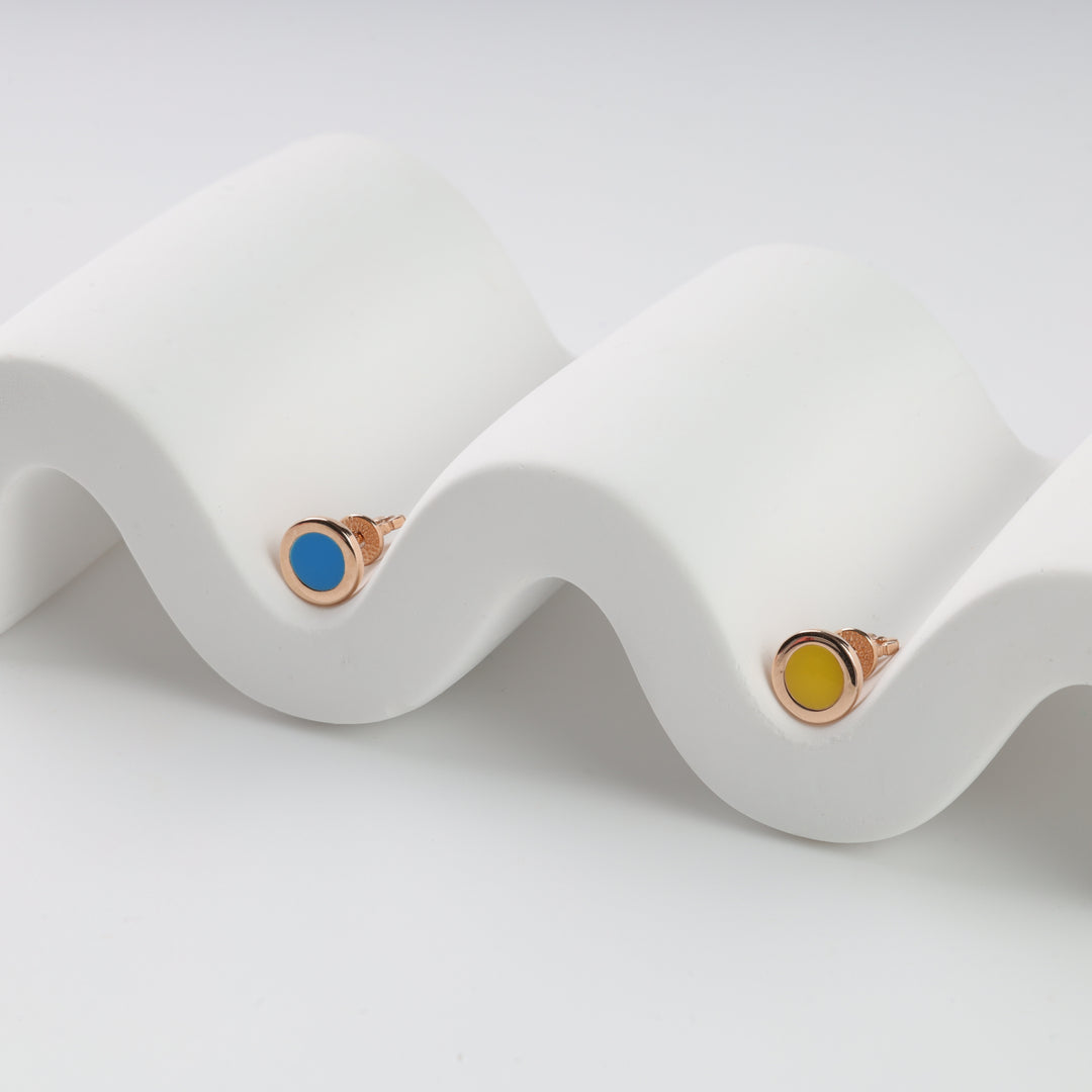 Two round stud earrings with gold settings, one with a blue center and one yellow, displayed on a white wavy surface.