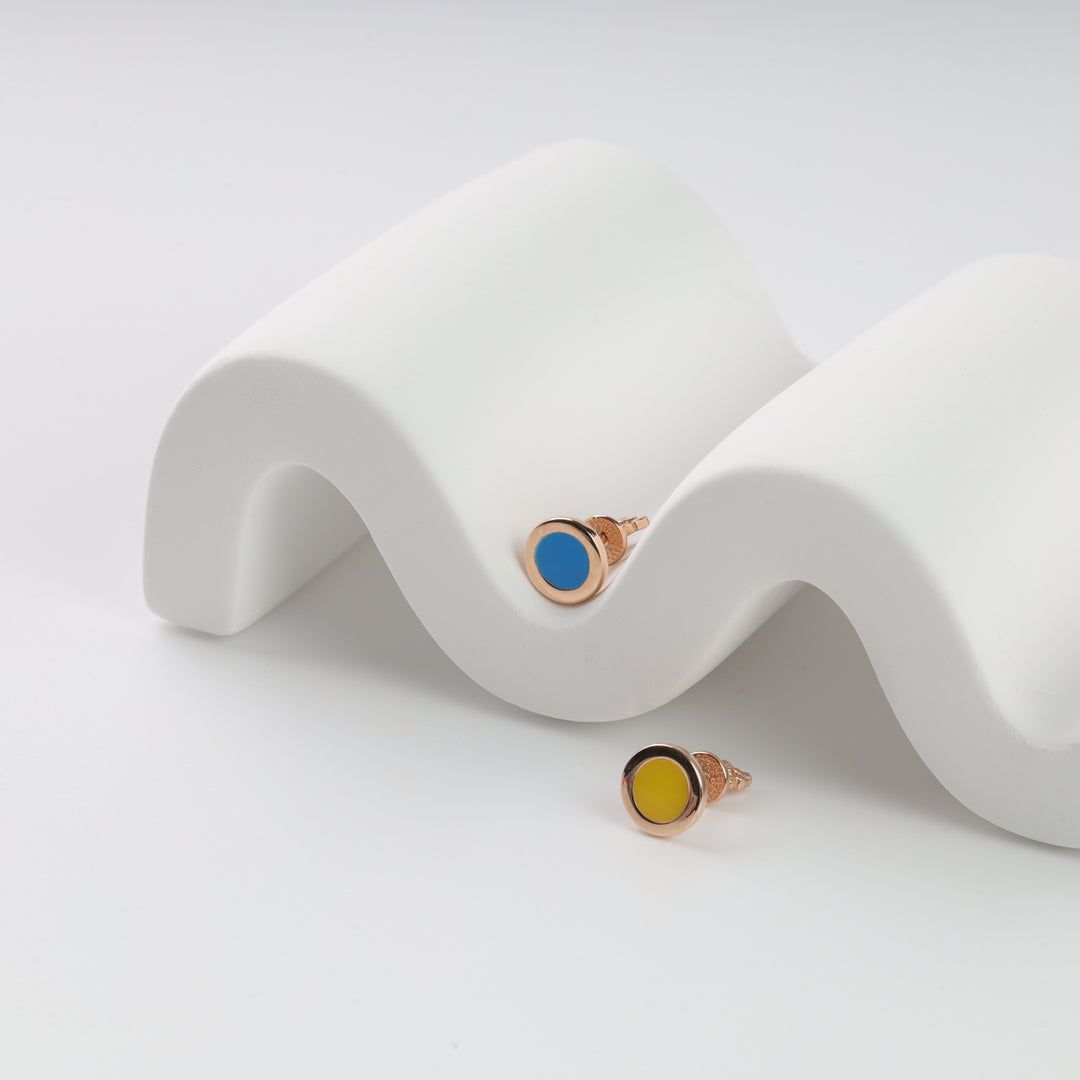 Pair of round stud earrings with gold settings, one with blue enamel and one with yellow enamel, displayed on a white wavy stand.