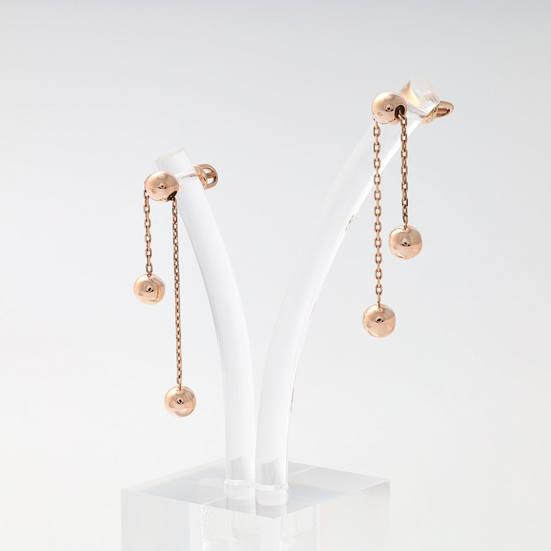 Pair of rose gold dangle earrings with three polished ball charms on delicate chains, displayed on a white stand.