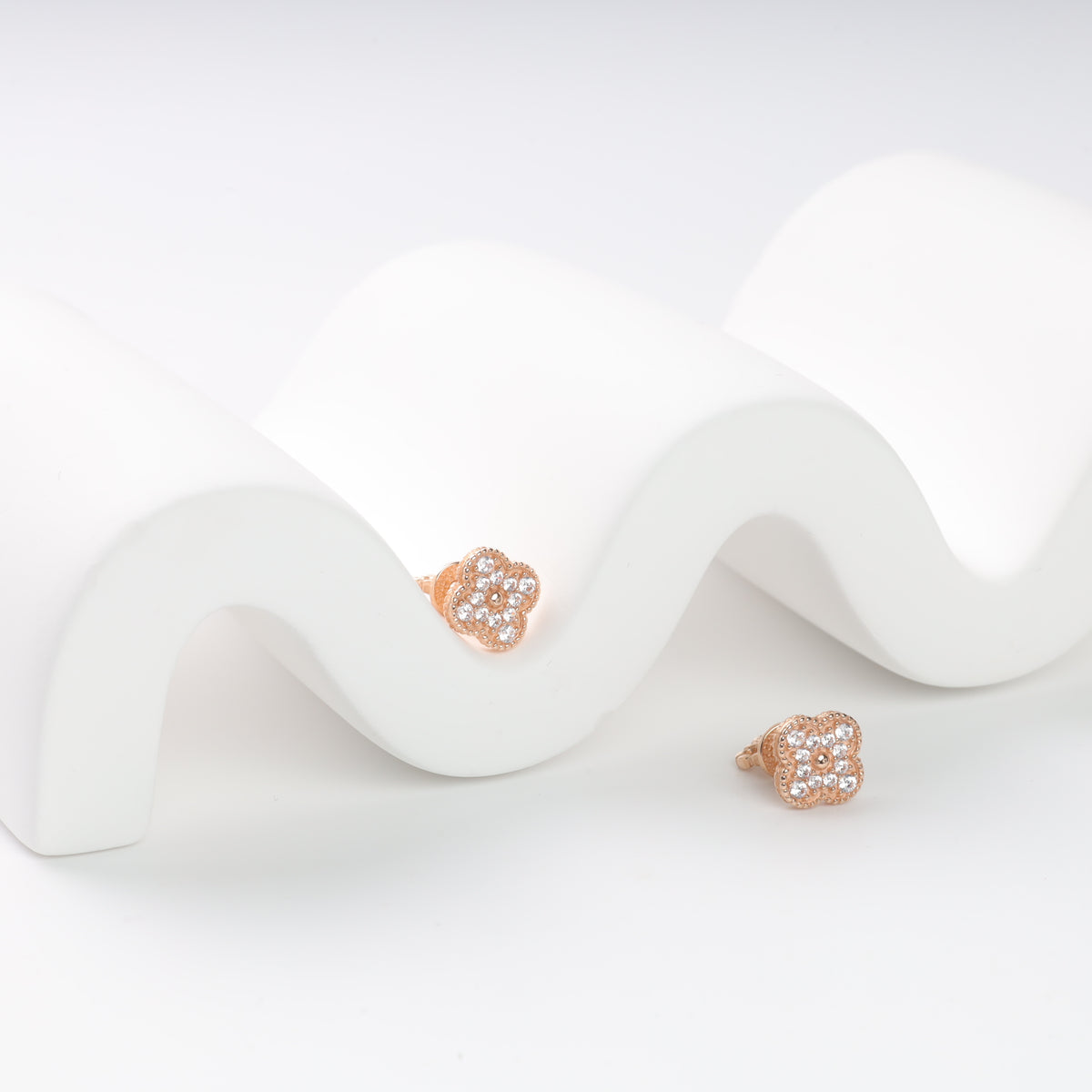 Rose gold flower-shaped stud earrings with clear crystals on a wavy white surface.