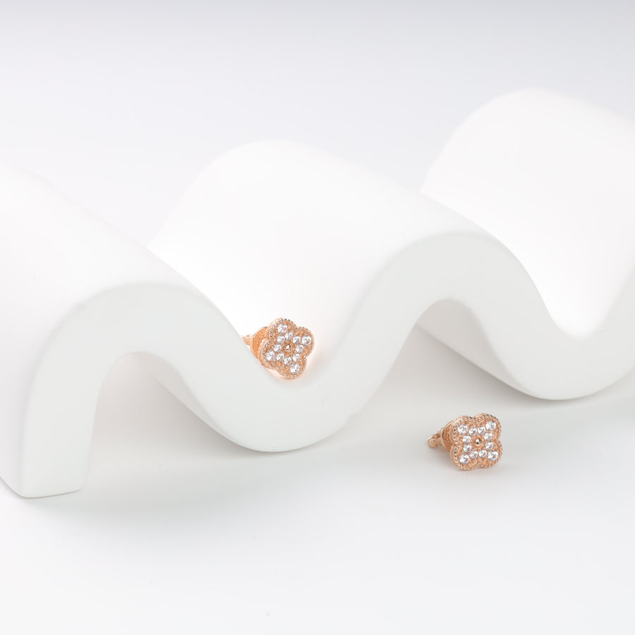 Rose gold flower-shaped stud earrings with clear crystals on a wavy white surface.
