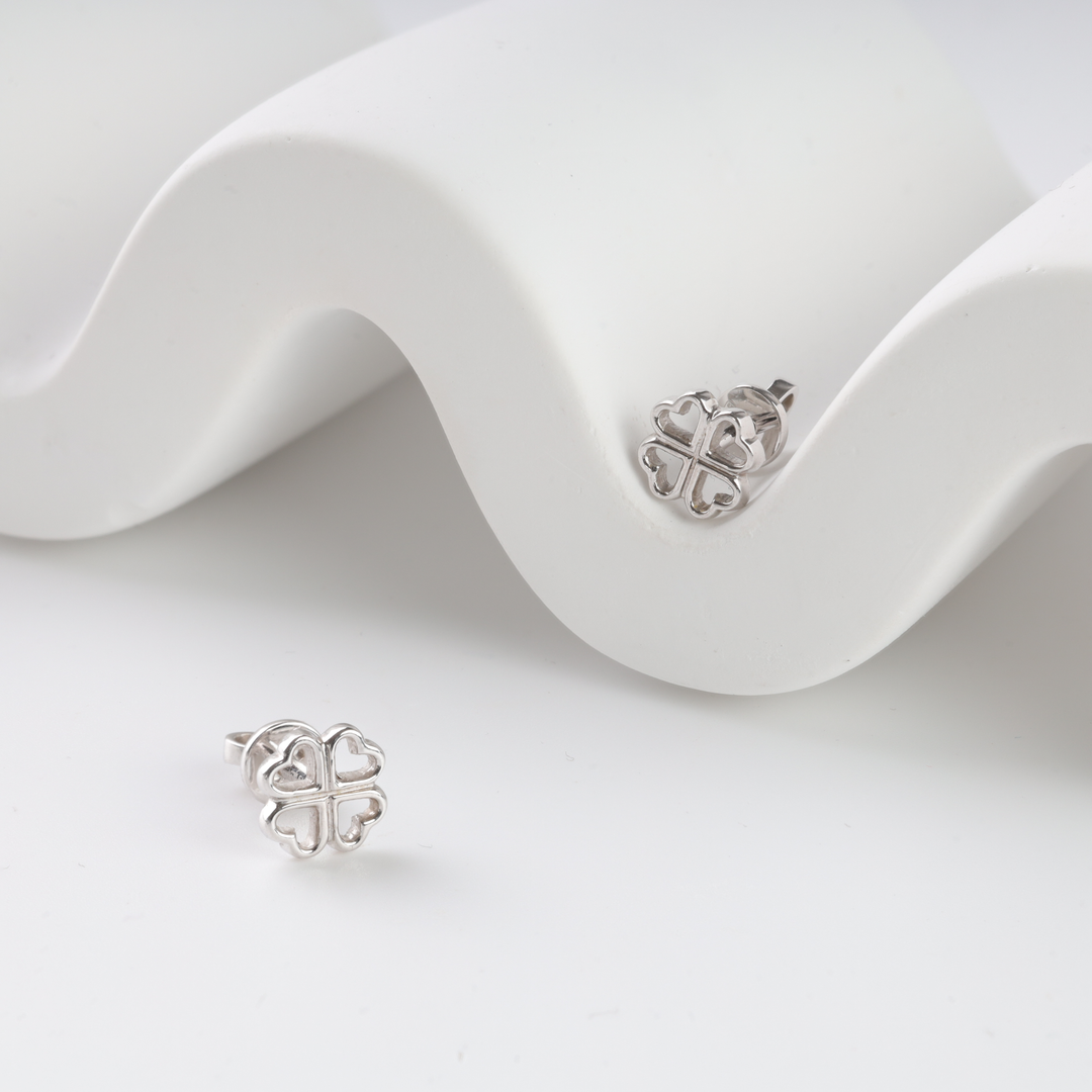Pair of silver stud earrings shaped like four-leaf clovers with heart petals on a white wavy display. Silver stud earrings, clover.