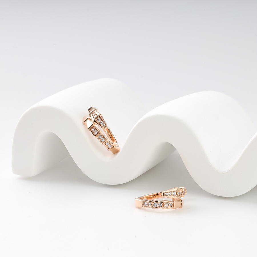 Pair of rose gold huggie hoop earrings with geometric design and small clear crystals displayed on white wavy stand.