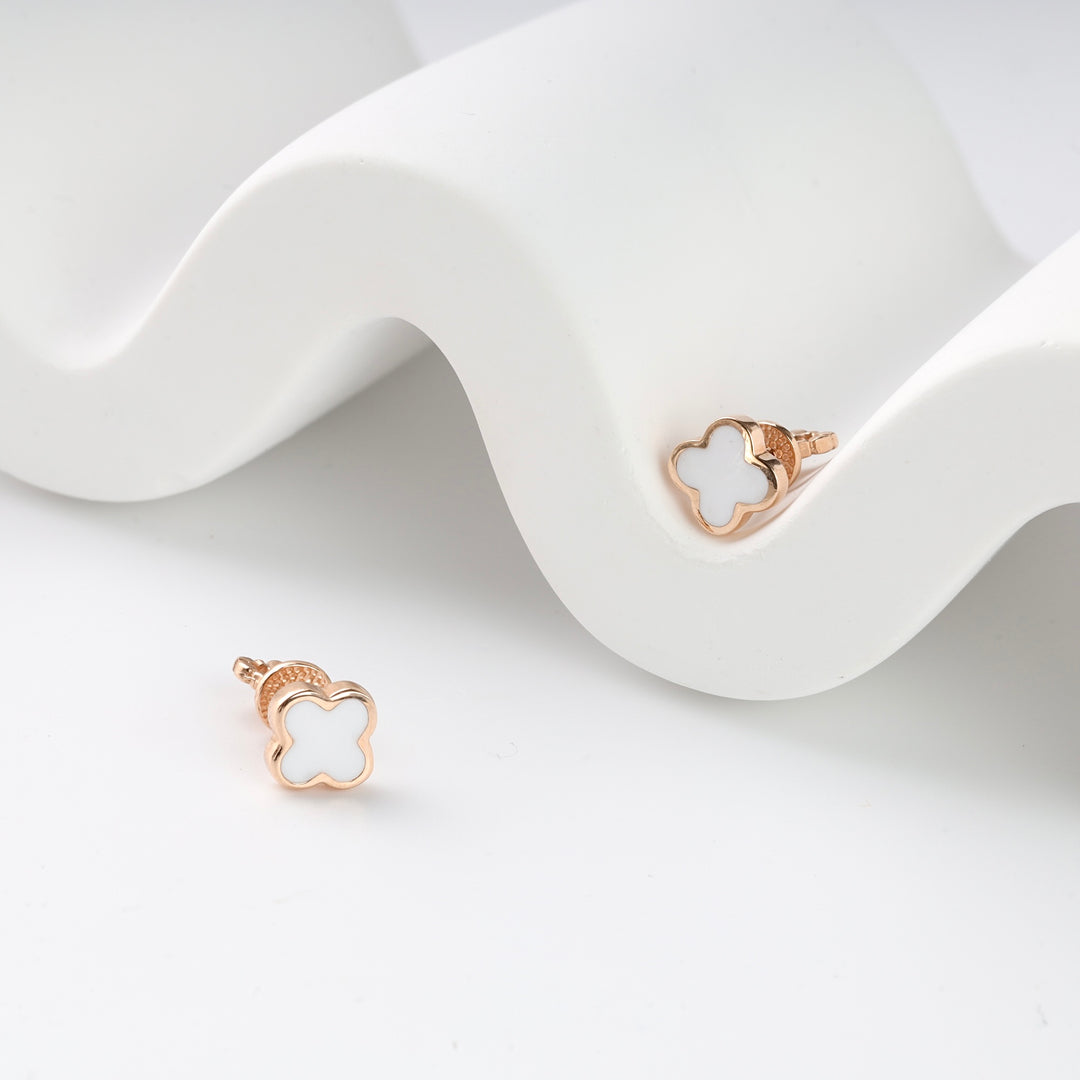 Pair of gold clover stud earrings with white enamel inlay displayed on a white wavy surface.
