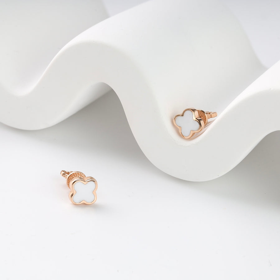 Pair of gold clover stud earrings with white enamel inlay displayed on a white wavy surface.