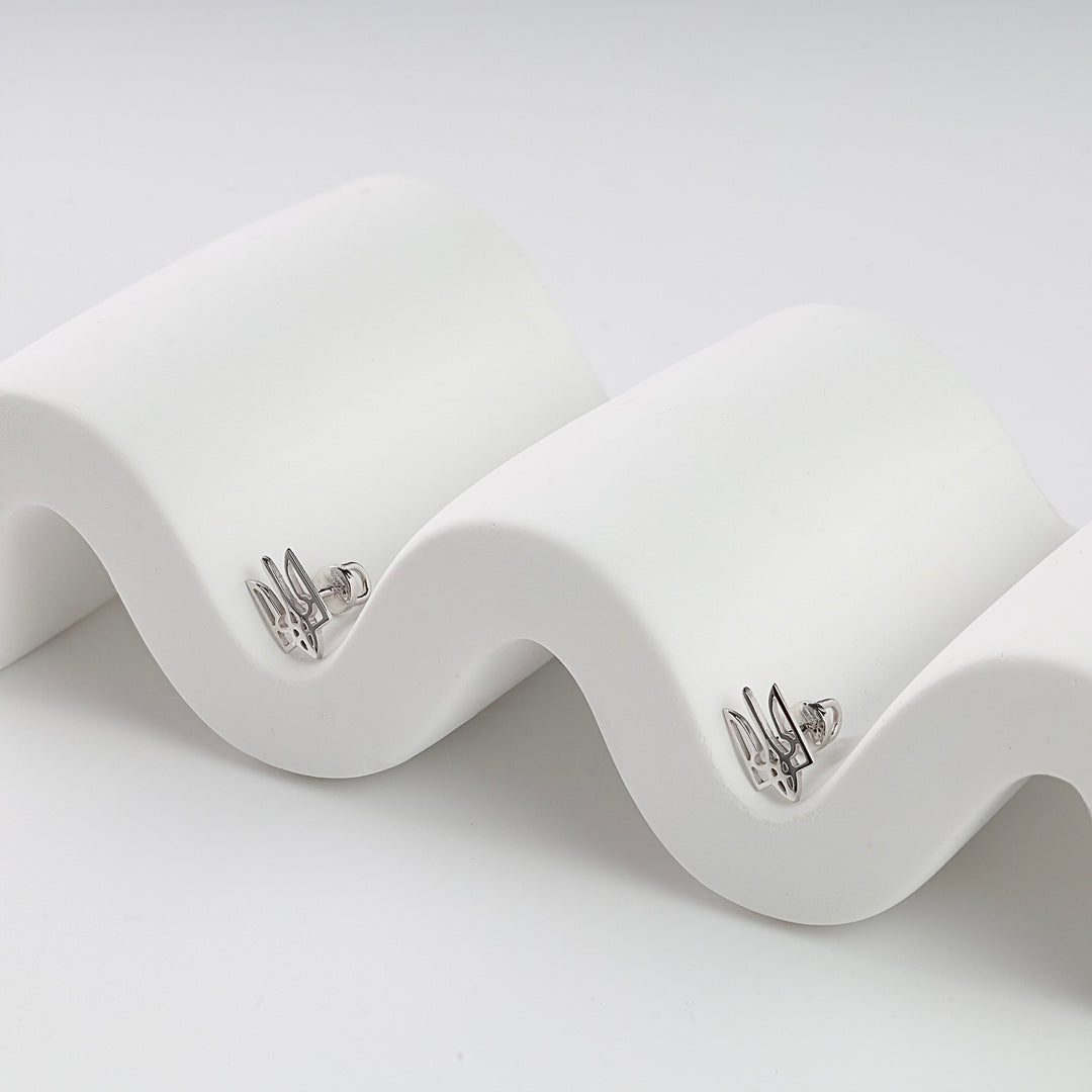 Pair of silver stud earrings with Ukrainian trident motif displayed on a white wavy stand.