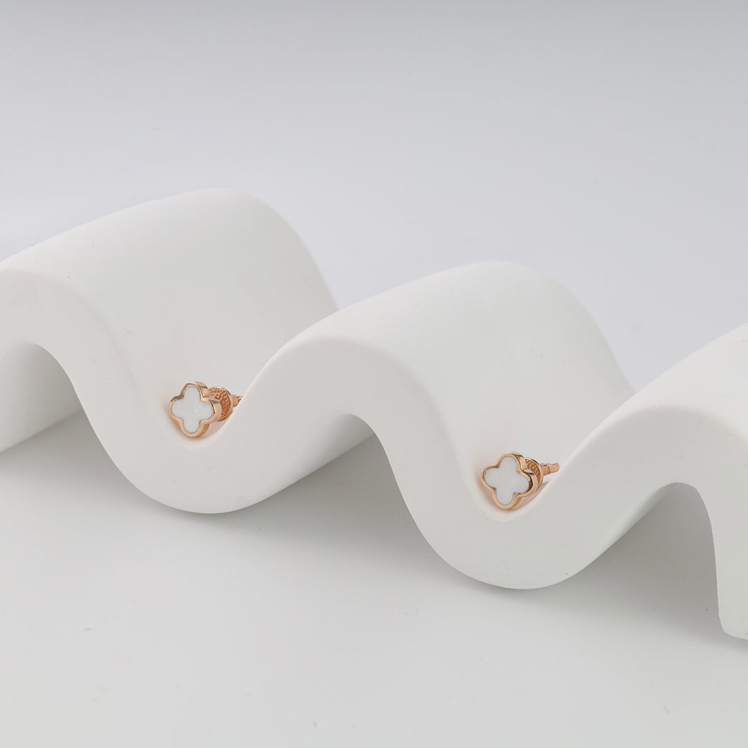 Pair of gold stud earrings with white clover shape, displayed on a wavy white stand, minimalist jewelry, stud earrings.