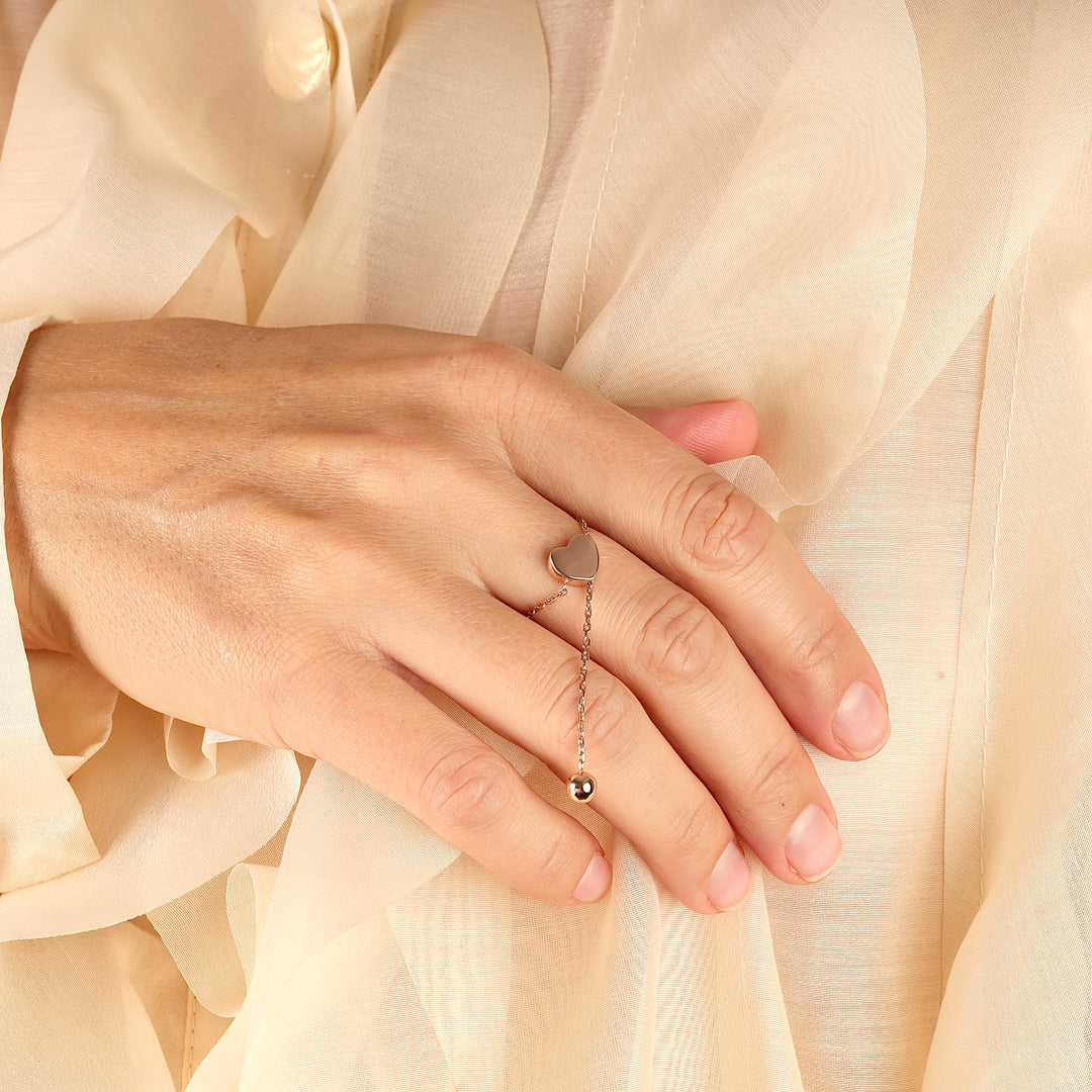Silver heart chain ring with dangling bead charm worn on a hand against a beige sheer fabric background.