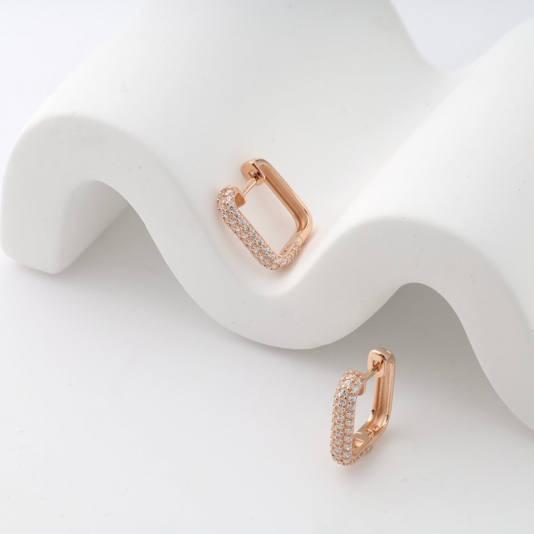 Rose gold rectangular hoop earrings with small crystals displayed on a white wavy surface.