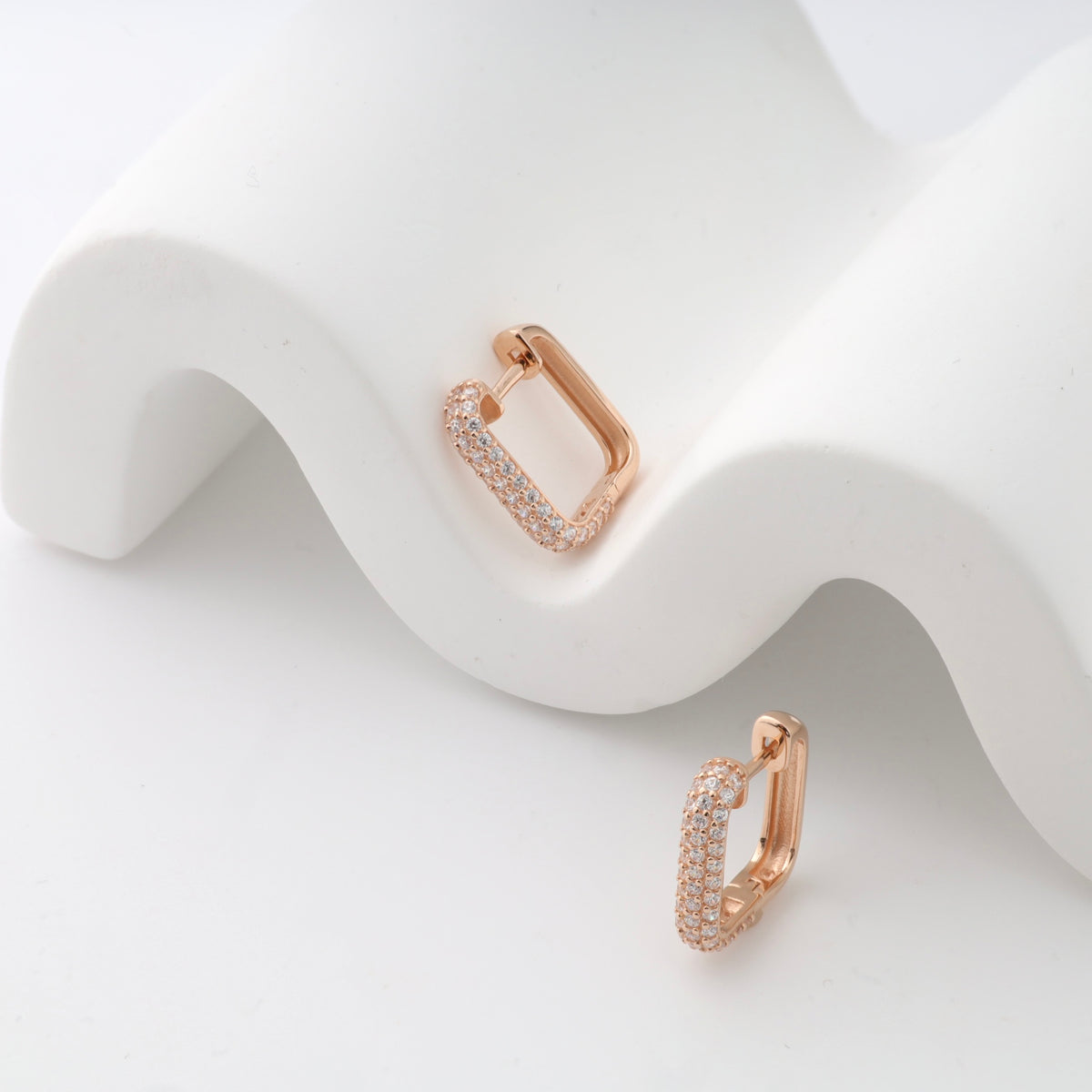 Rose gold rectangular hoop earrings with small crystals displayed on a white wavy surface.