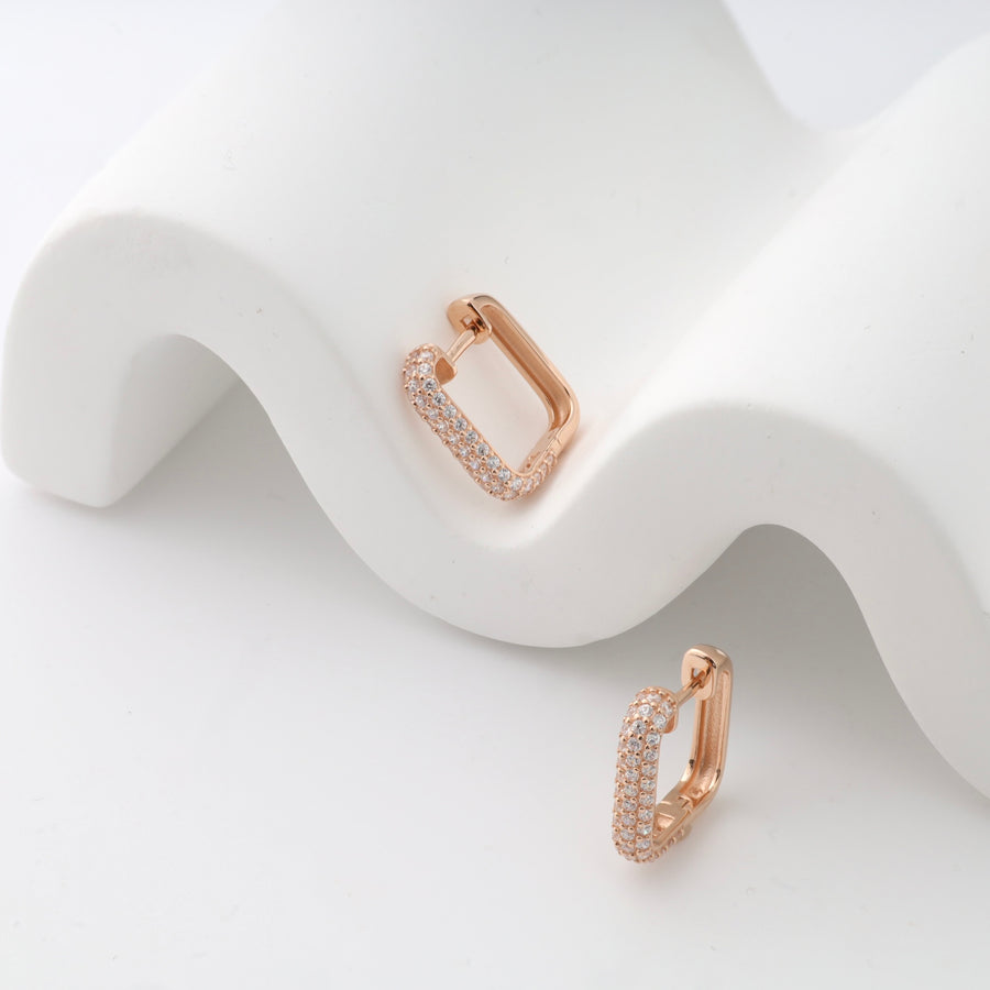 Rose gold rectangular hoop earrings with small crystals displayed on a white wavy surface.