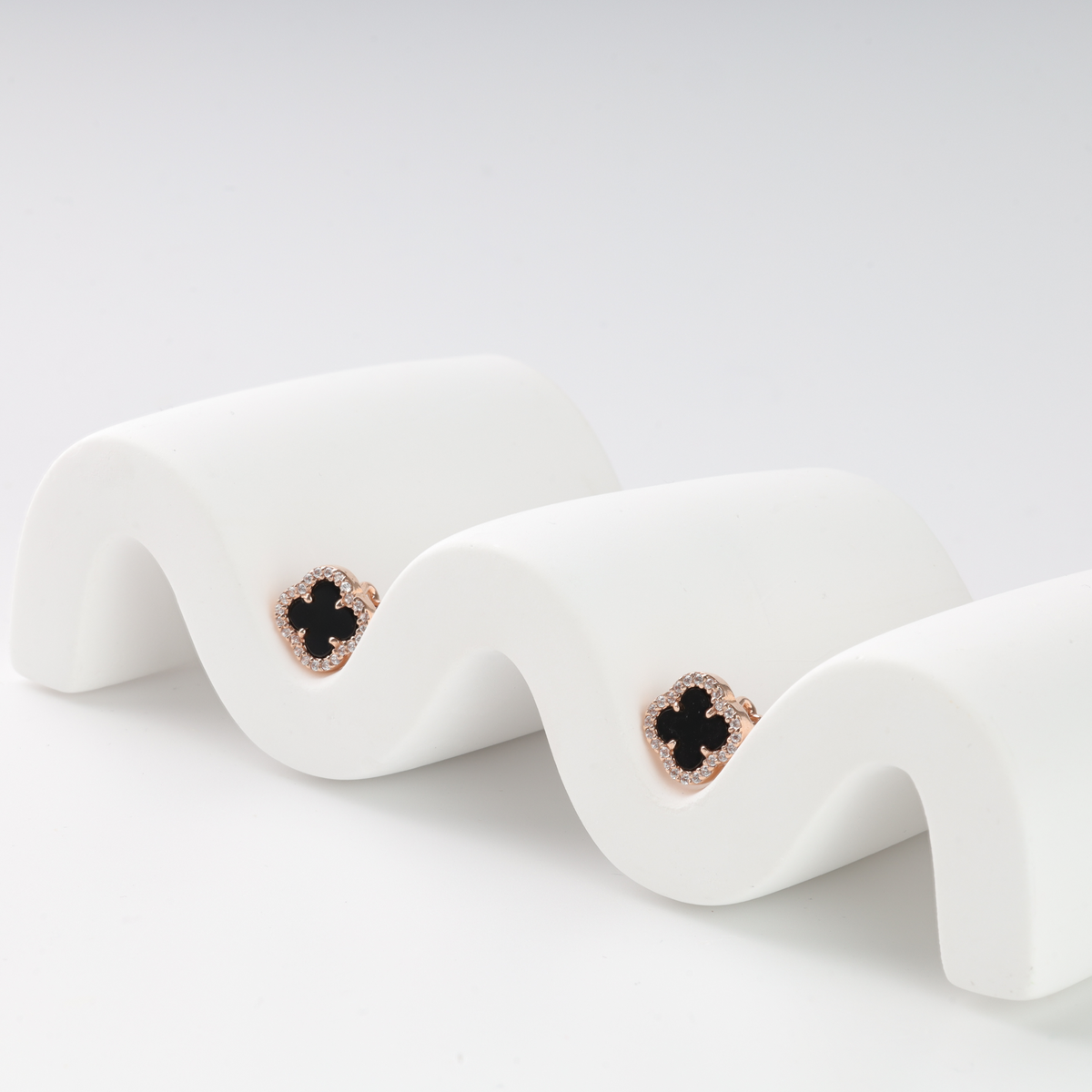 Pair of rose gold clover stud earrings with black stone and small crystals displayed on a white wavy stand.