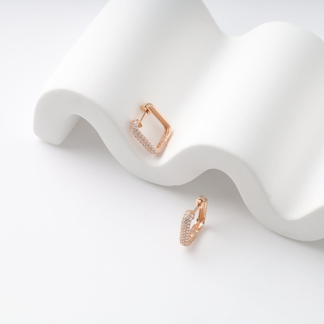 Two rose gold pavé huggie earrings on a wavy white surface.