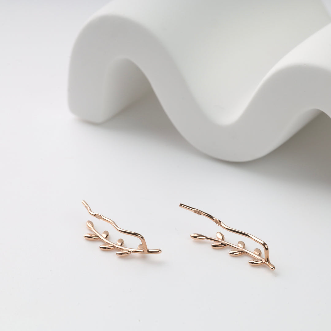 Pair of rose gold leaf ear climber earrings on a white surface beside a wavy display prop, ear climbers and leaf earrings.