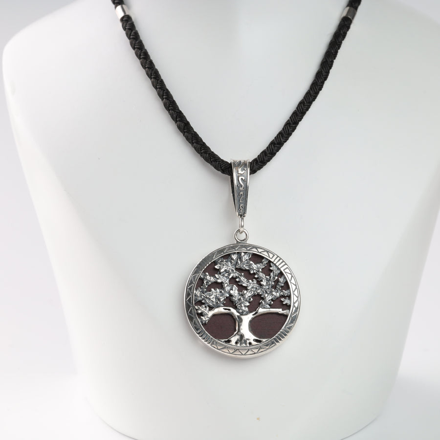 Silver tree of life pendant on a black braided necklace displayed on a white stand.