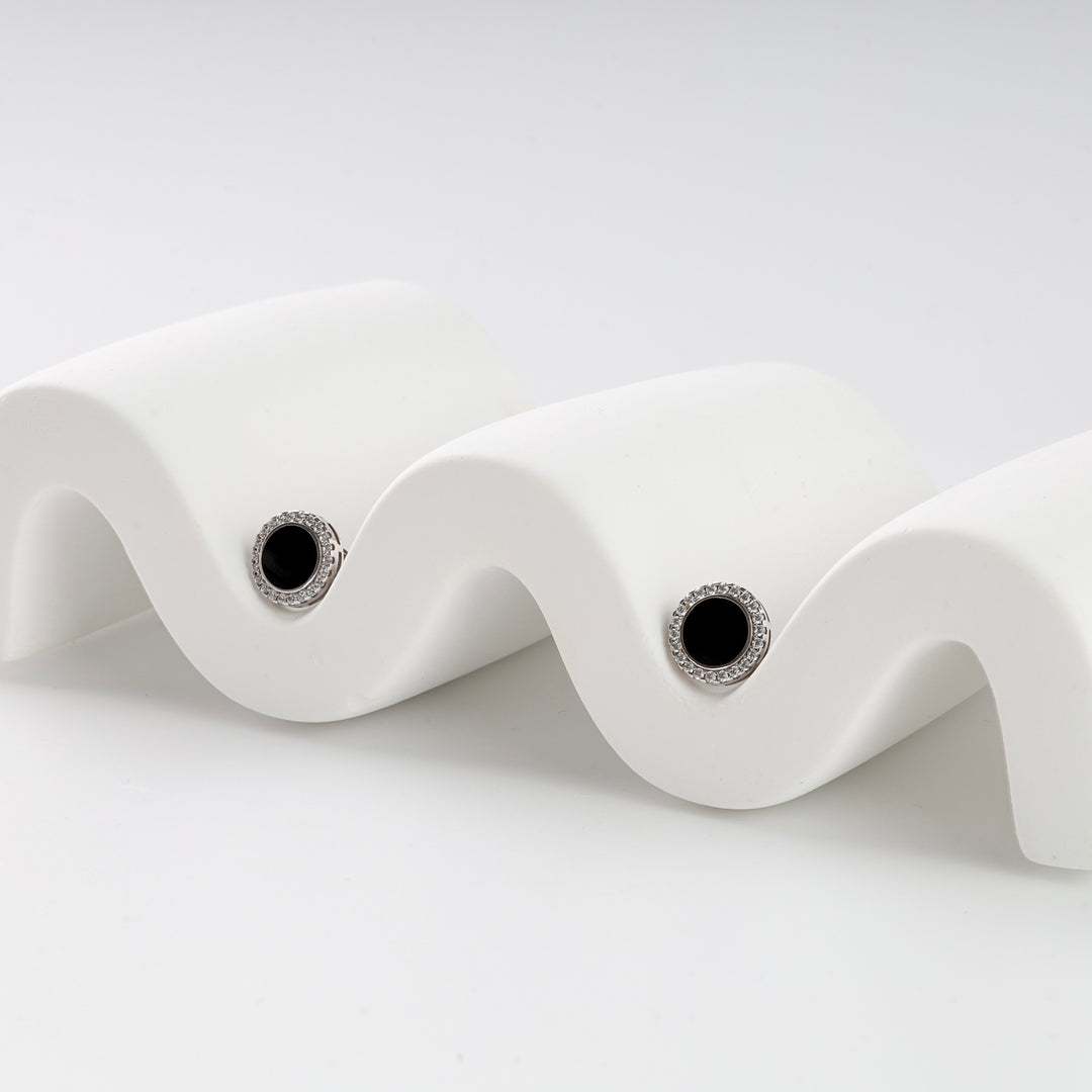 Pair of round black onyx stud earrings with silver halo setting on a white wavy display, gemstone stud earrings.