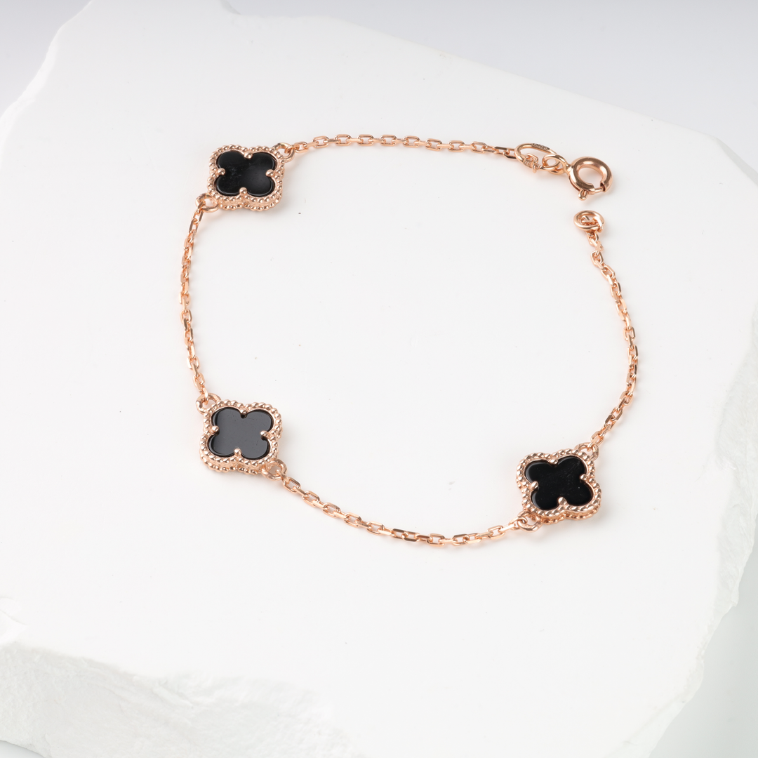 Gold chain bracelet with three black clover-shaped charms displayed on a white stone background.